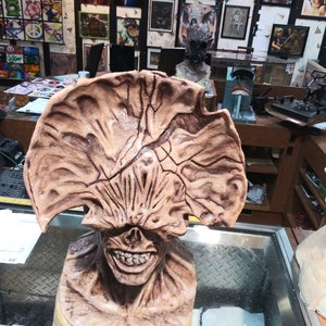 May include: A detailed, hand-painted, brown and beige sculpture of a creature with a large, open mouth and a single eye. The sculpture is displayed on a glass surface.