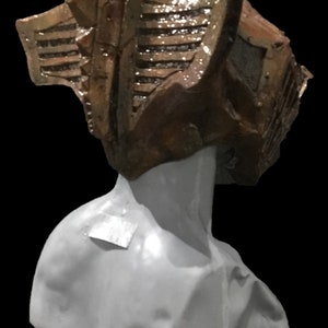 May include: A sculpture of a bust with a textured, brown headpiece. The headpiece has a geometric design with horizontal lines and a shiny finish. The bust is a light gray color, set against a black background.