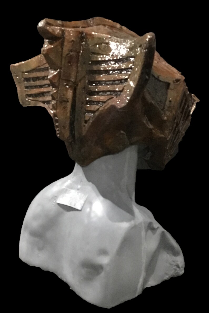 May include: A sculpture of a bust with a textured, brown headpiece. The headpiece has a geometric design with horizontal lines and a shiny finish. The bust is a light gray color, set against a black background.