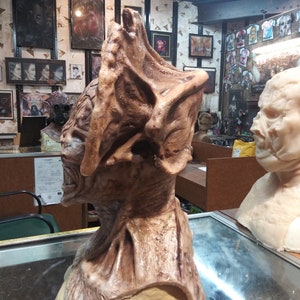 May include: A detailed, brown and beige, sculpted, monster mask with a large, open mouth and sharp teeth. The mask is on a beige stand.