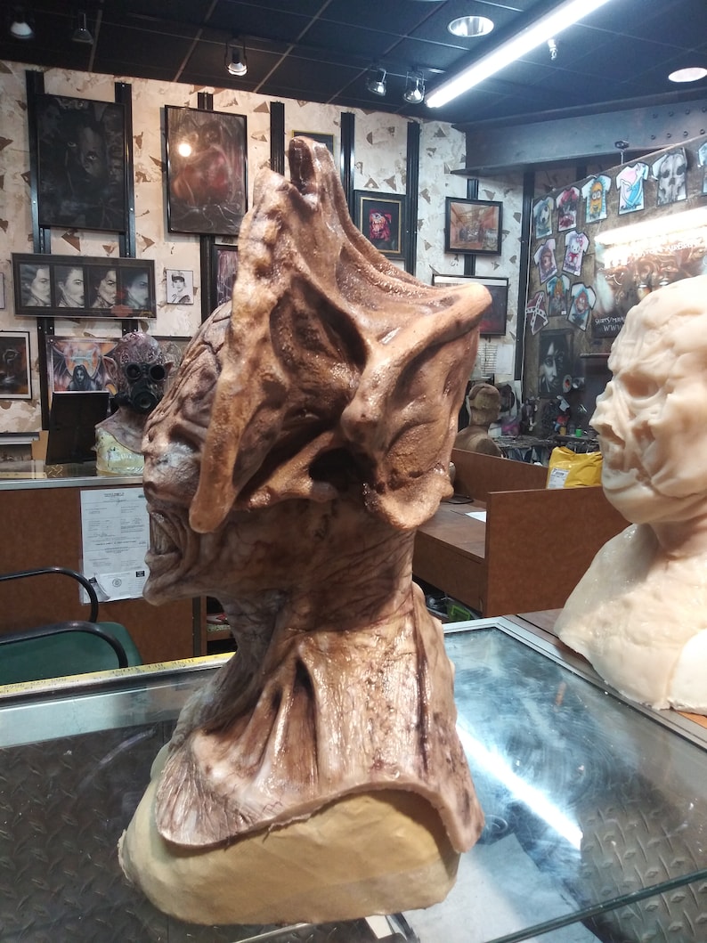 May include: A detailed, brown and beige, sculpted, monster mask with a large, open mouth and sharp teeth. The mask is on a beige stand.