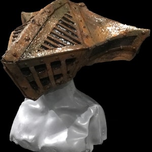 May include: A weathered, metallic helmet with a cage-like design and riveted details, resting on a white form. The helmet features a bronze and brown color scheme, suggesting an aged or antique appearance. The background is solid black.