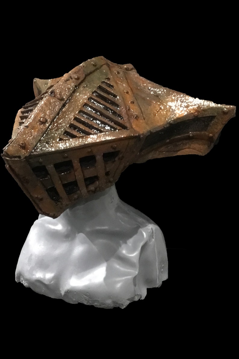 May include: A weathered, metallic helmet with a cage-like design and riveted details, resting on a white form. The helmet features a bronze and brown color scheme, suggesting an aged or antique appearance. The background is solid black.