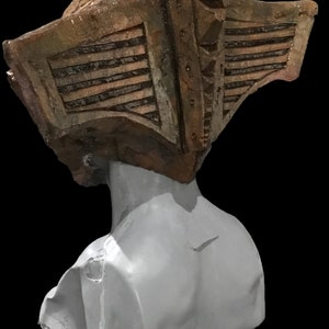 May include: A sculpture of a bust with a weathered, textured headpiece. The headpiece is a mix of brown and tan, with horizontal lines and rivet details. The bust itself is a smooth, light gray color, set against a black background.