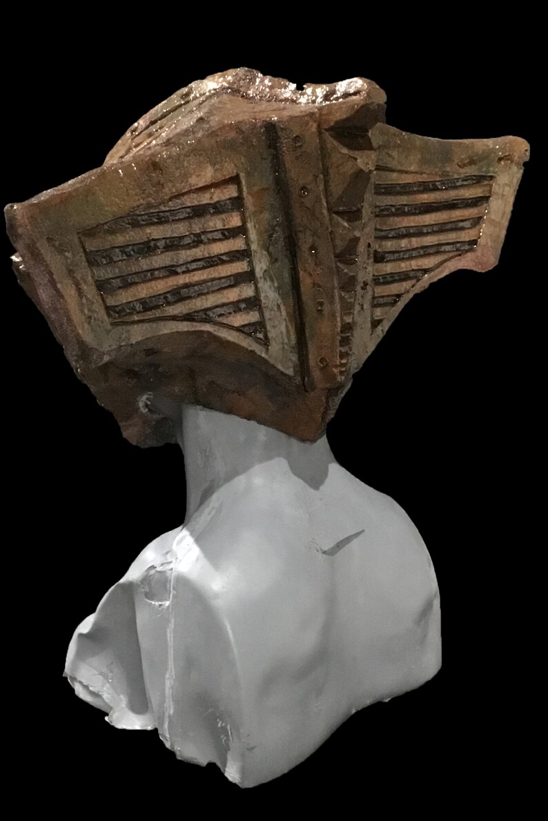 May include: A sculpture of a bust with a weathered, textured headpiece. The headpiece is a mix of brown and tan, with horizontal lines and rivet details. The bust itself is a smooth, light gray color, set against a black background.