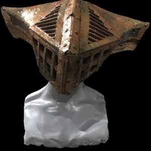 May include: A weathered, metallic sculpture of a helmet with a cage-like design. The helmet is a mix of brown and bronze tones, with visible rivets and a textured surface. The sculpture is mounted on a gray, abstract form.