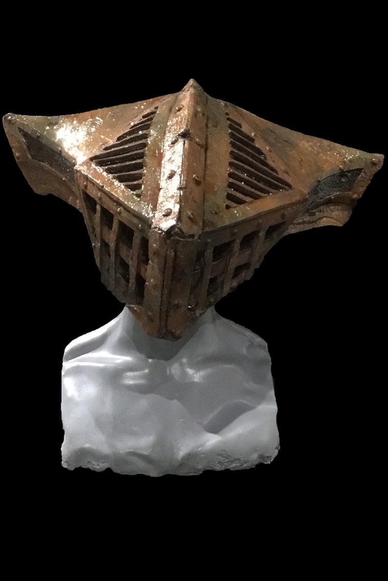 May include: A weathered, metallic sculpture of a helmet with a cage-like design. The helmet is a mix of brown and bronze tones, with visible rivets and a textured surface. The sculpture is mounted on a gray, abstract form.