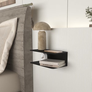 Può includere: Una mensola a muro nera a due livelli. Il ripiano superiore contiene uno smartphone e una piccola lampada. Il ripiano inferiore mostra due libri. Le mensole sono montate su una parete bianca a righe verticali, vicino a un letto.