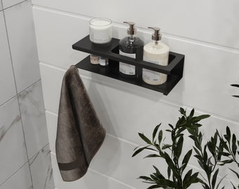 Può includere: Una mensola da bagno nera a due livelli con una candela, due dispenser di sapone e piccole bottiglie. Un asciugamano marrone è appeso sotto. La mensola è montata su una parete bianca con pannelli orizzontali. Pianta verde fogliosa nell'angolo.