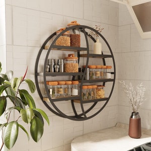 Puede incluir: Una estantería circular de metal negro con varios niveles, que contiene frascos de vidrio con diversas especias y hierbas. La estantería está montada en una pared de azulejos blancos.