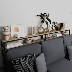 Op de afbeelding: Een modern, tweelaags schap met een lichte houten afwerking en een zwart metalen frame, geplaatst boven een grijze bank. De planken tonen decoratieve vazen, boeken, ingelijste kunst en een plant in een glazen vaas.