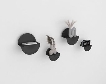 May include: Four black half-circle wall shelves with rounded edges. The shelves are mounted on a white wall. The top shelf has a small gray vase with a gray abstract sculpture. The second shelf has a small gray vase with a small plant. The third shelf has a cell phone. The bottom shelf has a small black digital clock that reads "08:15".