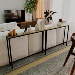 Może przedstawiać: Czarny metalowy stolik konsolowy z drewnianym blatem, na którym stoi biała ceramiczna wazon, czarna figurka ptaka i stos książek. Stolik stoi przed białą sofą i dywanem.