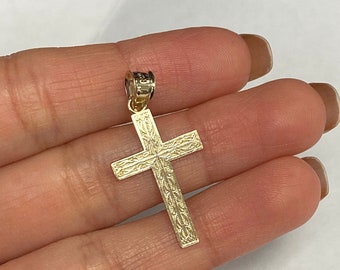 Puede incluir: Un colgante de cruz de color dorado con un diseño grabado ornamental. La cruz presenta un patrón detallado en sus brazos y centro, y un pequeño lazo para una cadena. El colgante se sostiene en una mano sobre un fondo blanco.