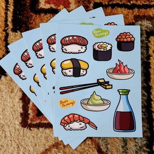 May include: A stack of light blue stickers featuring colorful sushi illustrations. The stickers showcase various sushi types, including nigiri, maki rolls, and a bottle of soy sauce. The text "Reda Printings" is visible on the stickers.