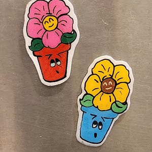 Könnte beinhalten: Zwei Topfblumenmagnete mit fröhlichen Gesichtern. Die rosa Blume hat ein gelbes Gesicht mit einem Lächeln und die gelbe Blume hat ein braunes Gesicht mit einem Lächeln. Die Töpfe haben blaue und rote Gesichter mit besorgten Ausdrücken.