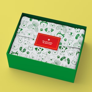 Pode incluir: Uma caixa de presente quadrada verde contendo papel de seda branco estampado com rostos de cães e pegadas em verde. Uma etiqueta retangular vermelha com borda dourada apresenta um ícone de pata e o texto For pawrents, by pawrents.