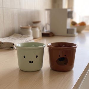 Pode incluir: Duas canecas de cerâmica com bordas arredondadas. A caneca da esquerda é verde-clara e apresenta um gráfico preto simples de dois olhos e uma boca em forma de X. A caneca da direita é marrom e exibe um gráfico de rosto de urso.