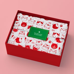 Può includere: Una scatola regalo rossa contenente carta velina bianca con un motivo ripetuto di teste di gatto rosse e la parola MEOW. Un adesivo rettangolare verde con bordo dorato è centrato sulla carta, con un'impronta di zampa e il testo For pawrents, by pawrents.