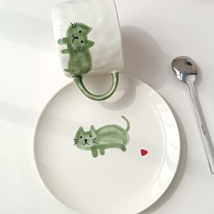 Può includere: Una tazza in ceramica bianca con superficie testurizzata e un'illustrazione di un gatto verde, abbinata a un piattino bianco con un gatto verde e un piccolo cuore rosso. Un cucchiaio in metallo argentato è posizionato a destra del piatto.