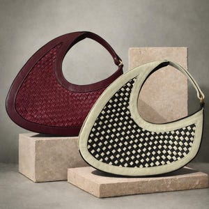 May include: Two woven leather handbags. One is a deep burgundy color, and the other is black and white. Both have a crescent shape with a handle at the top. They are displayed on small, square, stone blocks against a neutral background.