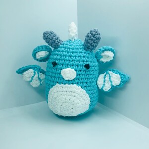 May include: A handmade crochet dragon in shades of blue, white, and grey. The amigurumi dragon has a rounded body, small wings, and a white horn. The dragon's belly and face are white, with black eyes.