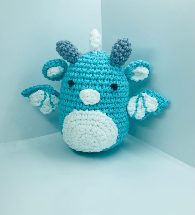 May include: A handmade crochet dragon in shades of blue, white, and grey. The amigurumi dragon has a rounded body, small wings, and a white horn. The dragon's belly and face are white, with black eyes.