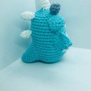 May include: A handmade crochet creature in shades of blue, white, and grey. The plush toy has a rounded body with white cloud-like accents and a grey pom-pom on top. The crochet stitches are visible, giving it a textured appearance.