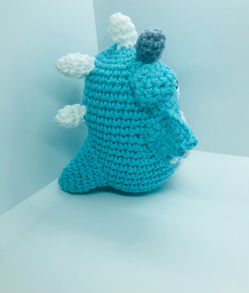 May include: A handmade crochet creature in shades of blue, white, and grey. The plush toy has a rounded body with white cloud-like accents and a grey pom-pom on top. The crochet stitches are visible, giving it a textured appearance.