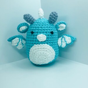 May include: A handmade crochet dragon in shades of blue, white, and grey. The dragon has small wings, ears, and a horn. The body is a light blue with a white belly. The wings and ears have white accents. The dragon is a whimsical, soft toy.