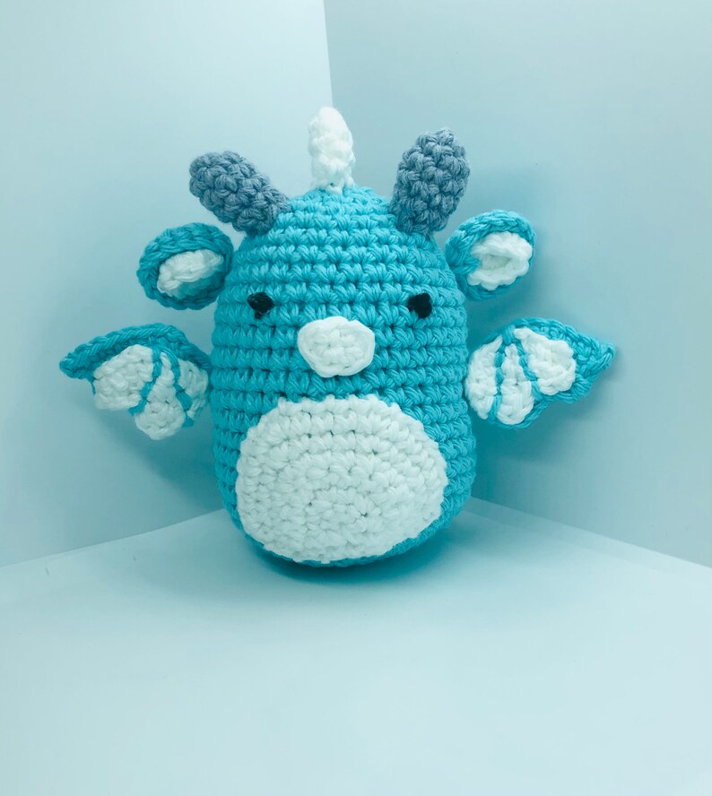 May include: A handmade crochet dragon in shades of blue, white, and grey. The dragon has small wings, ears, and a horn. The body is a light blue with a white belly. The wings and ears have white accents. The dragon is a whimsical, soft toy.