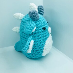 May include: A handmade crochet dinosaur toy in shades of blue, white, and grey. The toy features a rounded body, small black eyes, and decorative spikes. The dinosaur is approximately 15 cm tall.