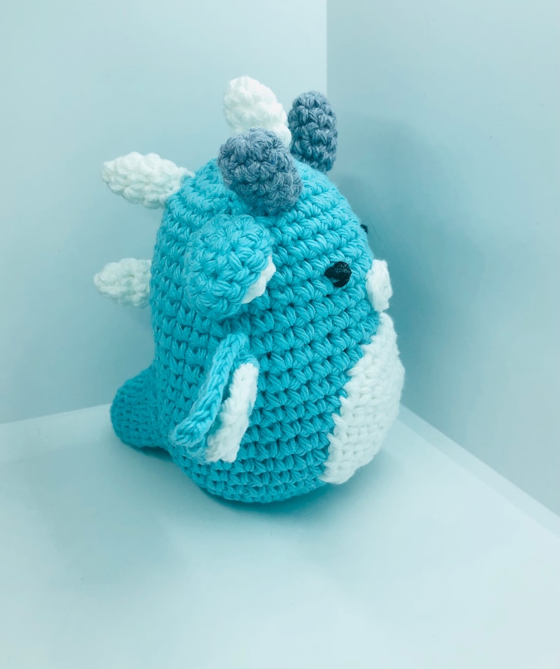 May include: A handmade crochet dinosaur toy in shades of blue, white, and grey. The toy features a rounded body, small black eyes, and decorative spikes. The dinosaur is approximately 15 cm tall.