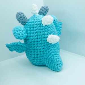May include: A handmade, light blue crochet creature with white and grey accents. The plush toy has a rounded body, small wings, and multiple protruding shapes on its head. The toy is made of soft yarn and is suitable for children.