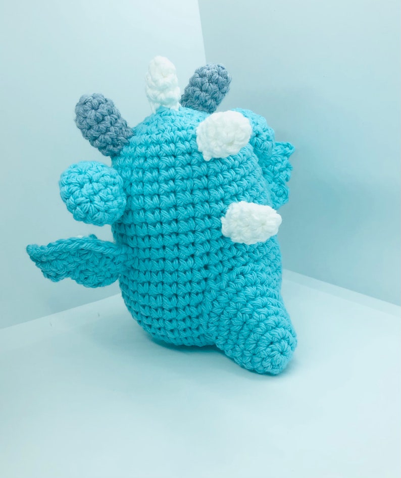 May include: A handmade, light blue crochet creature with white and grey accents. The plush toy has a rounded body, small wings, and multiple protruding shapes on its head. The toy is made of soft yarn and is suitable for children.