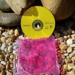 May include: A yellow CD-R audio disc with the text "SONY" and "CD-R AUDIO 80" printed on it. The disc is sitting on a bed of small, brown rocks. A pink CD case is sitting on the rocks in front of the yellow disc.