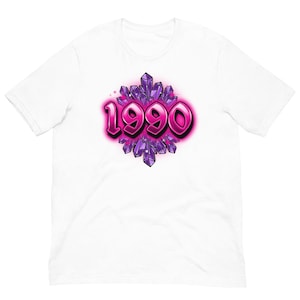 Peut inclure: T-shirt blanc avec un motif graphique. Le design présente le nombre "1990" dans une police rose, de style bulle, sur un fond de formes de cristaux violets. Le style rappelle les designs rétro ou vintage, avec une apparence audacieuse et accrocheuse.