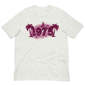 Könnte beinhalten: Hellgraues T-Shirt mit einem lilafarbenen Airbrush-Design der Zahl "1975", Palmen und einem Mond. Das Design hat einen Sternenhintergrund. Das T-Shirt ist ein kurzärmeliges Rundhals-Shirt.