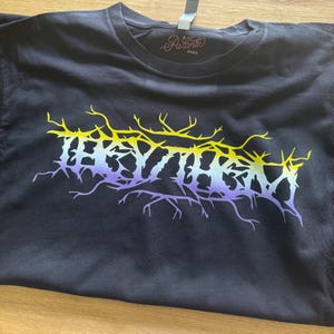 May include: Black t-shirt featuring a yellow, purple and white graphic of the word "RHYTHM" in a metal band font.