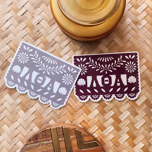 May include: Two paper cut-out style stickers with the word "ALOHA" in white lettering on a burgundy and a light gray background. The stickers have a traditional Mexican paper cut-out design with flowers and seashells.