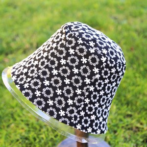May include: A black bucket hat with a white floral pattern. The hat is on a display stand.