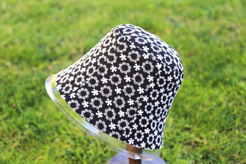 May include: A black bucket hat with a white floral pattern. The hat is on a display stand.