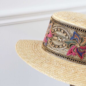 May include: A straw hat with a wide brim and a decorative band. The band features colorful floral embroidery with pink, blue, and gold accents, and is embellished with sequins. The hat is a light beige color.