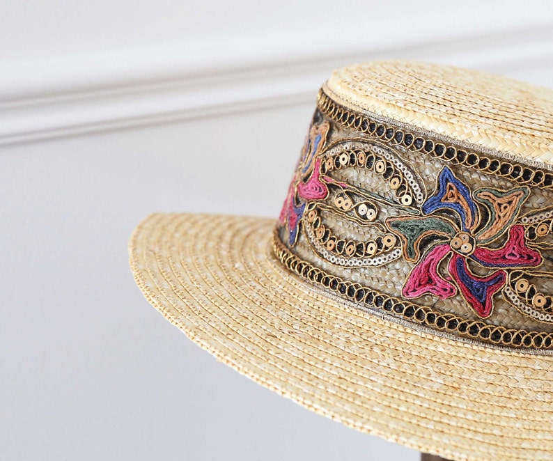 May include: A straw hat with a wide brim and a decorative band. The band features colorful floral embroidery with pink, blue, and gold accents, and is embellished with sequins. The hat is a light beige color.