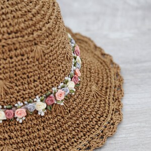 Puede incluir: Un sombrero de paja marr&oacute;n con un borde de ganchillo y una banda de flores de tela rosa, blanca y morada.