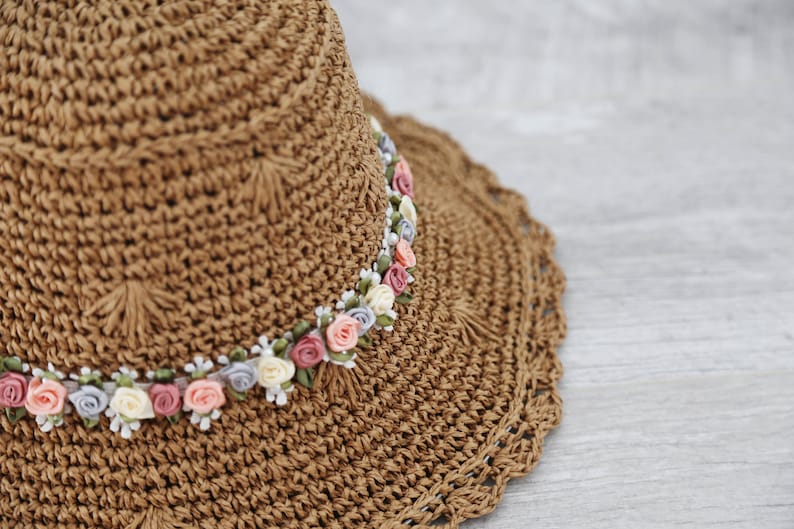 Puede incluir: Un sombrero de paja marr&oacute;n con un borde de ganchillo y una banda de flores de tela rosa, blanca y morada.