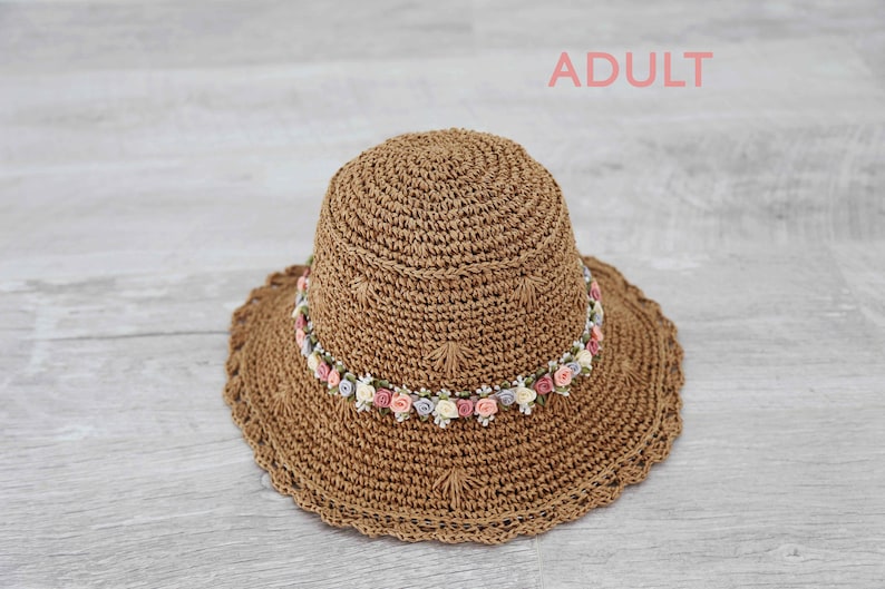 Puede incluir: Un sombrero de sol de crochet marr&oacute;n con una banda de flores rosas, blancas y azules.