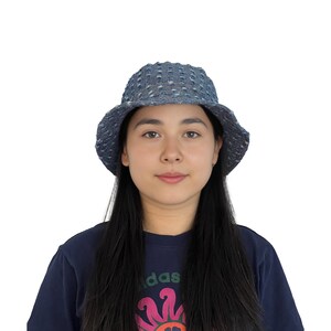 K&ouml;nnte beinhalten: Eine Person tr&auml;gt einen blauen Jeans-Bucket-Hut mit wei&szlig;em Muster. Der Hut hat eine breite Krempe und eine leicht abgerundete Krone. Die Person tr&auml;gt ein dunkelblaues Shirt mit einem bunten Grafikdesign.