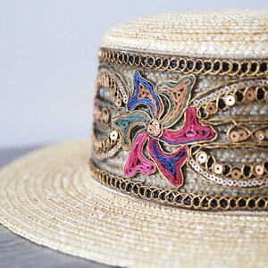 May include: A straw hat with a flat top and wide brim. The hat band is decorated with colorful embroidered floral designs, gold sequins, and black beads. The hat has a vintage or bohemian aesthetic.