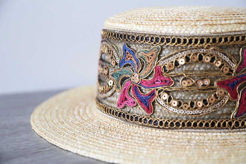 May include: A straw hat with a flat top and wide brim. The hat band is decorated with colorful embroidered floral designs, gold sequins, and black beads. The hat has a vintage or bohemian aesthetic.